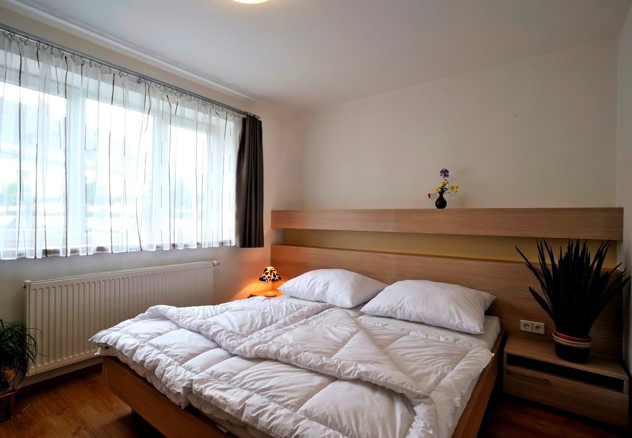 Modern eingerichtetes Schlafzimmer mit Doppelbett, hellem Holzmobiliar und großen Fenstern mit weißen Vorhängen.