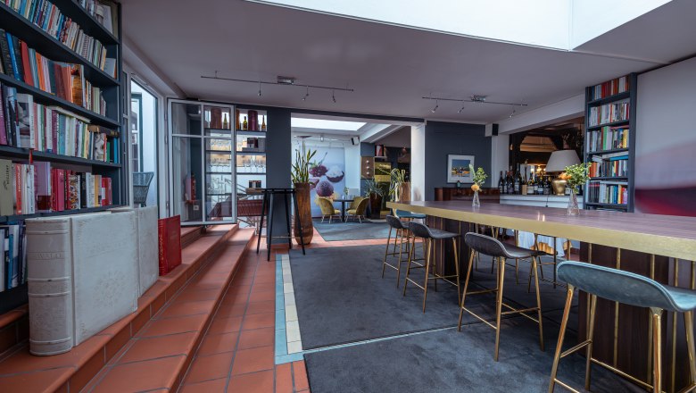 Innenansicht eines modernen Hotelraums mit Bücherregalen, hohen Tischen und Barhockern.