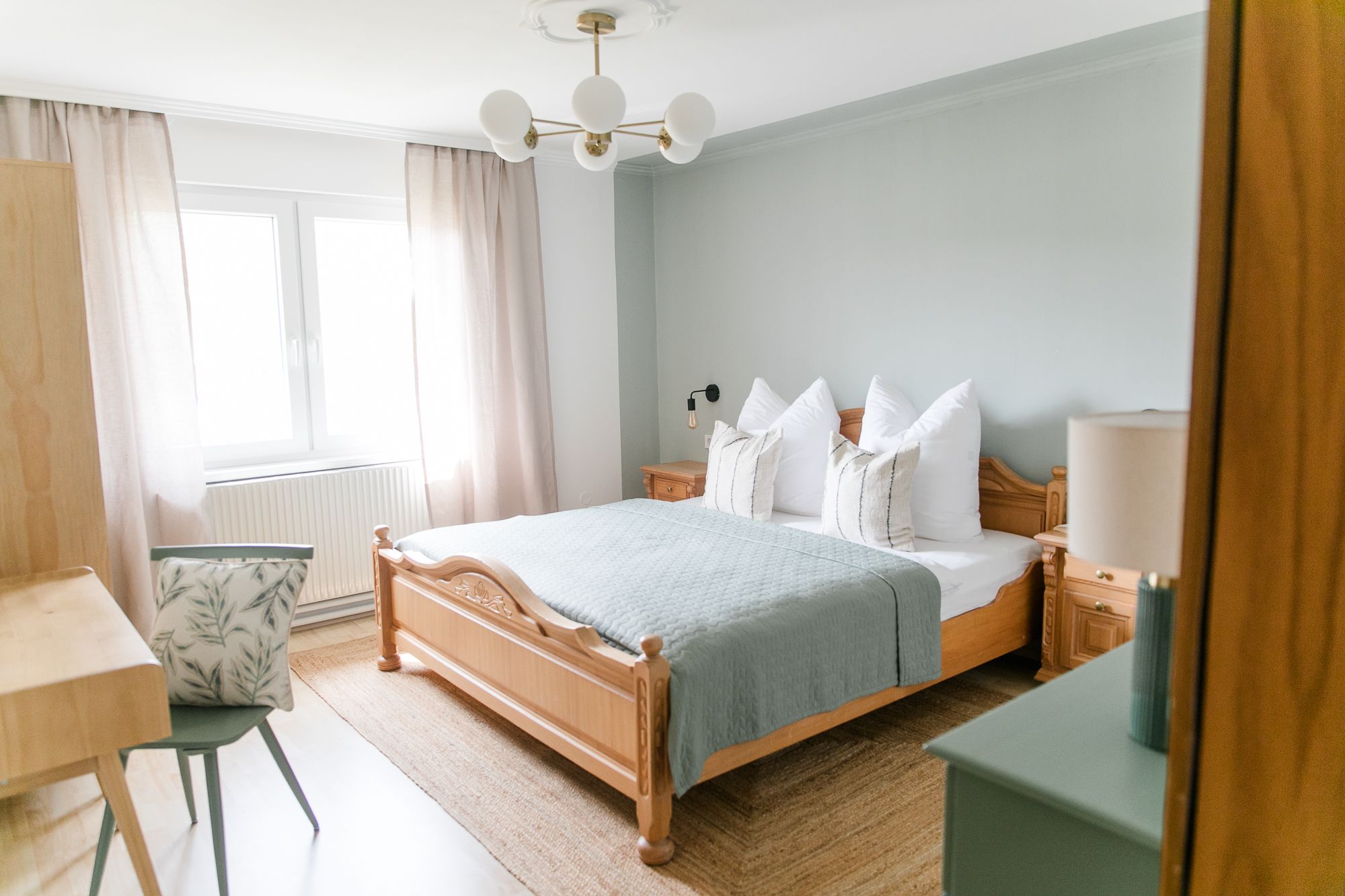 Ein gemütliches Schlafzimmer mit einem Holzbett, blauer Bettdecke, Nachttischen und einem Schreibtisch mit Stuhl. Helle Vorhänge und ein Fenster sind zu sehen.