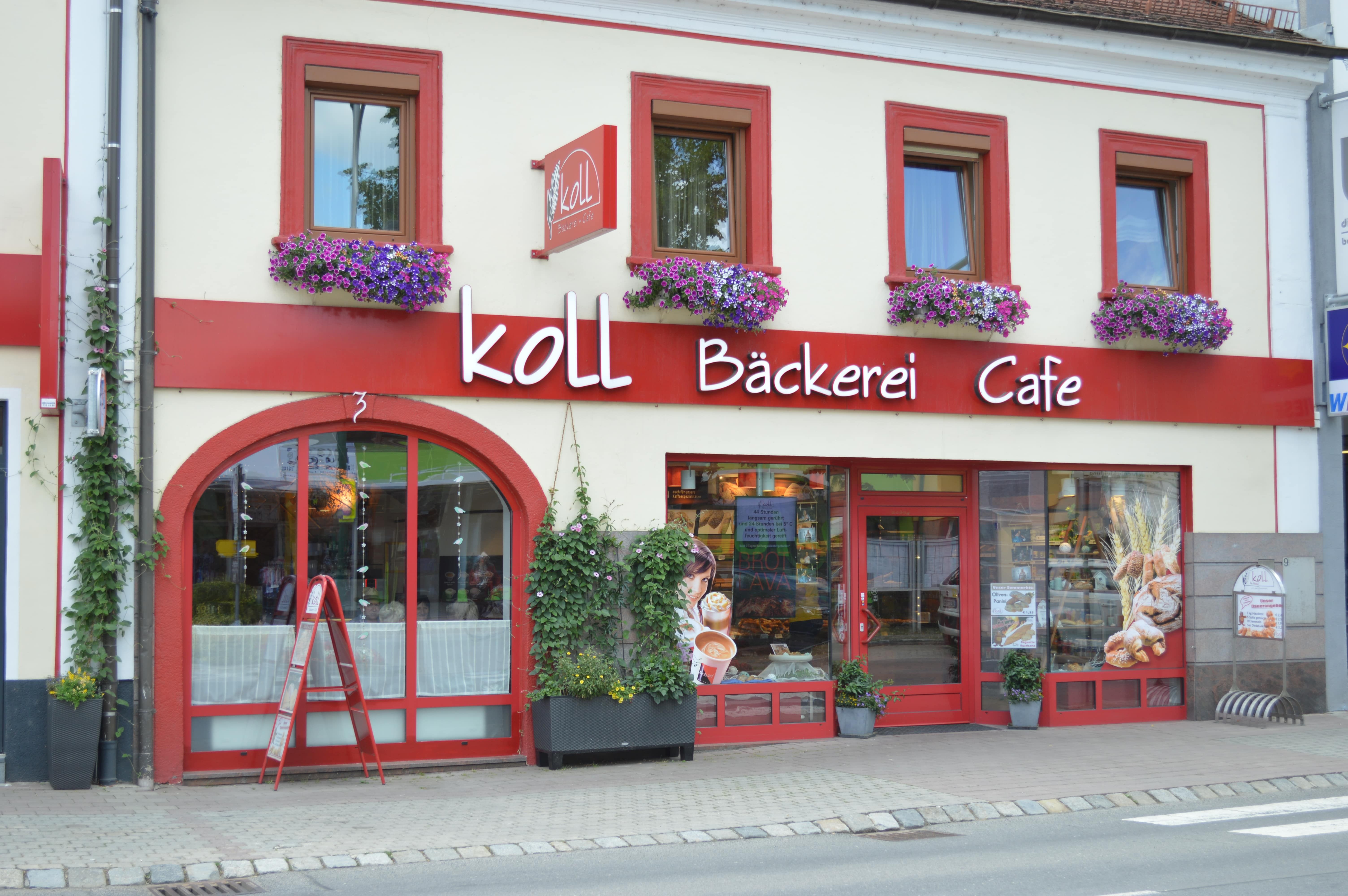 Außenansicht der Koll Bäckerei Café in Kirchschlag mit roten Fensterrahmen und Blumenkästen.