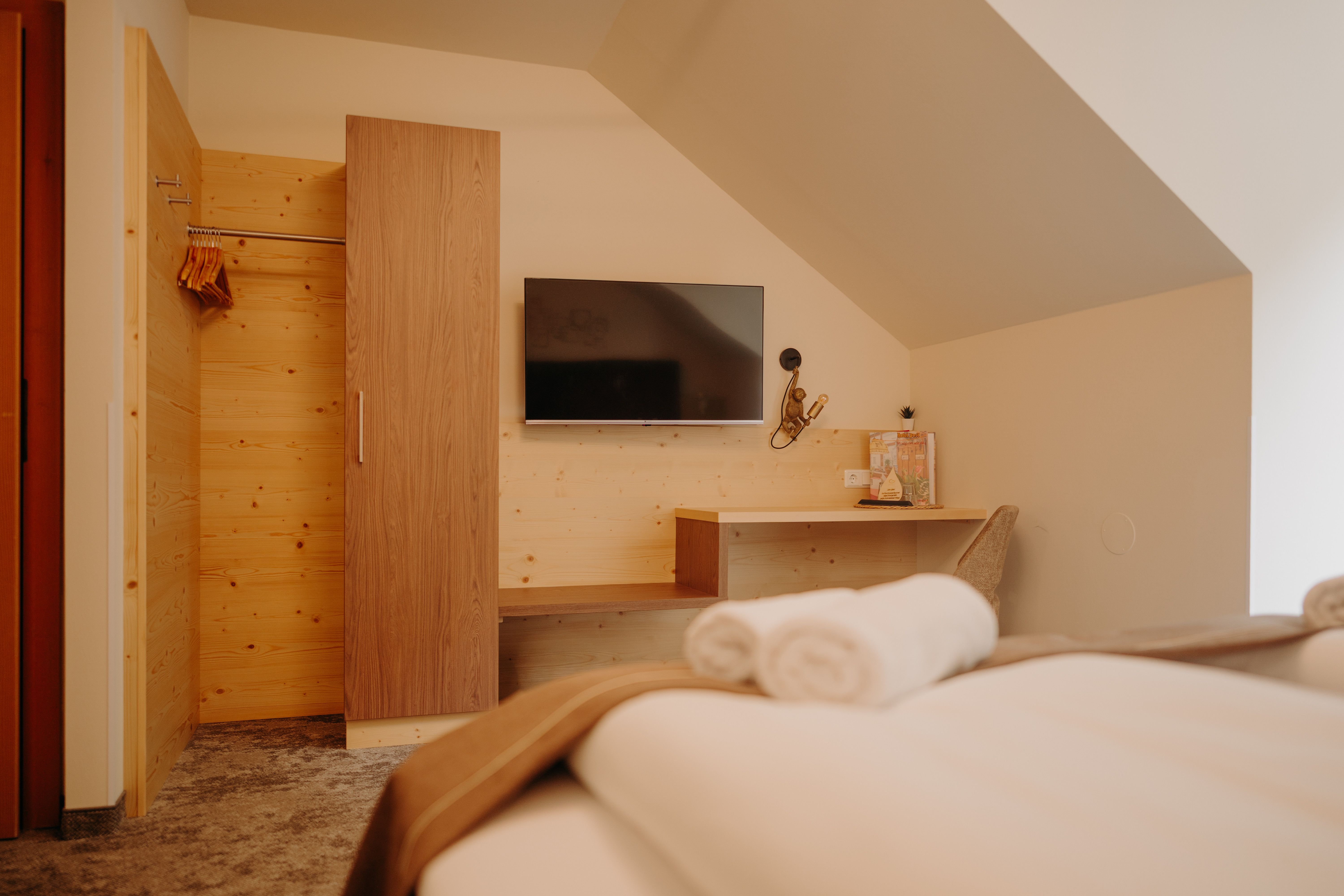 Ein gemütliches Hotelzimmer fotografiert vom Bett aus. Gegenüber vom Bett hängt ein Flachbildfernsehener an der Wand. Daneben ist eine Garderobe aus hellem Holz, die Wand ist auch zum Teil mit Holz verkleidet. Dort befindet sich auch eine Ablagefläche und ein Schreibtisch mit Sessel und Lampe. Das Zimmer hat eine Dachschräge.