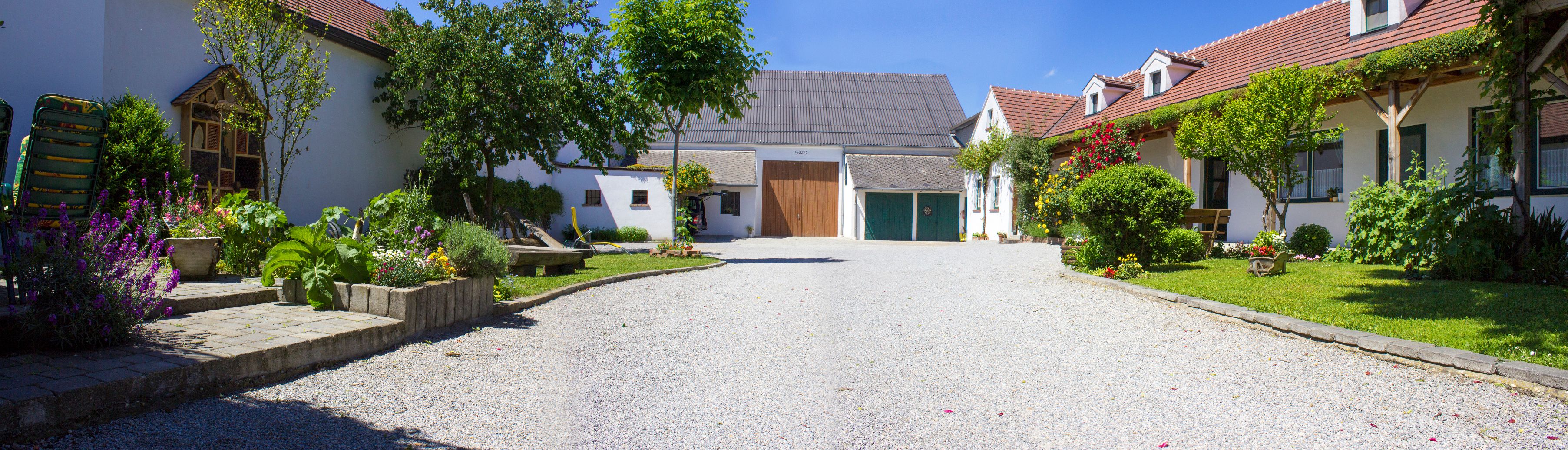 Ein idyllisches Weingut mit Kiesweg, blühenden Gärten und traditionellen Gebäuden unter blauem Himmel.