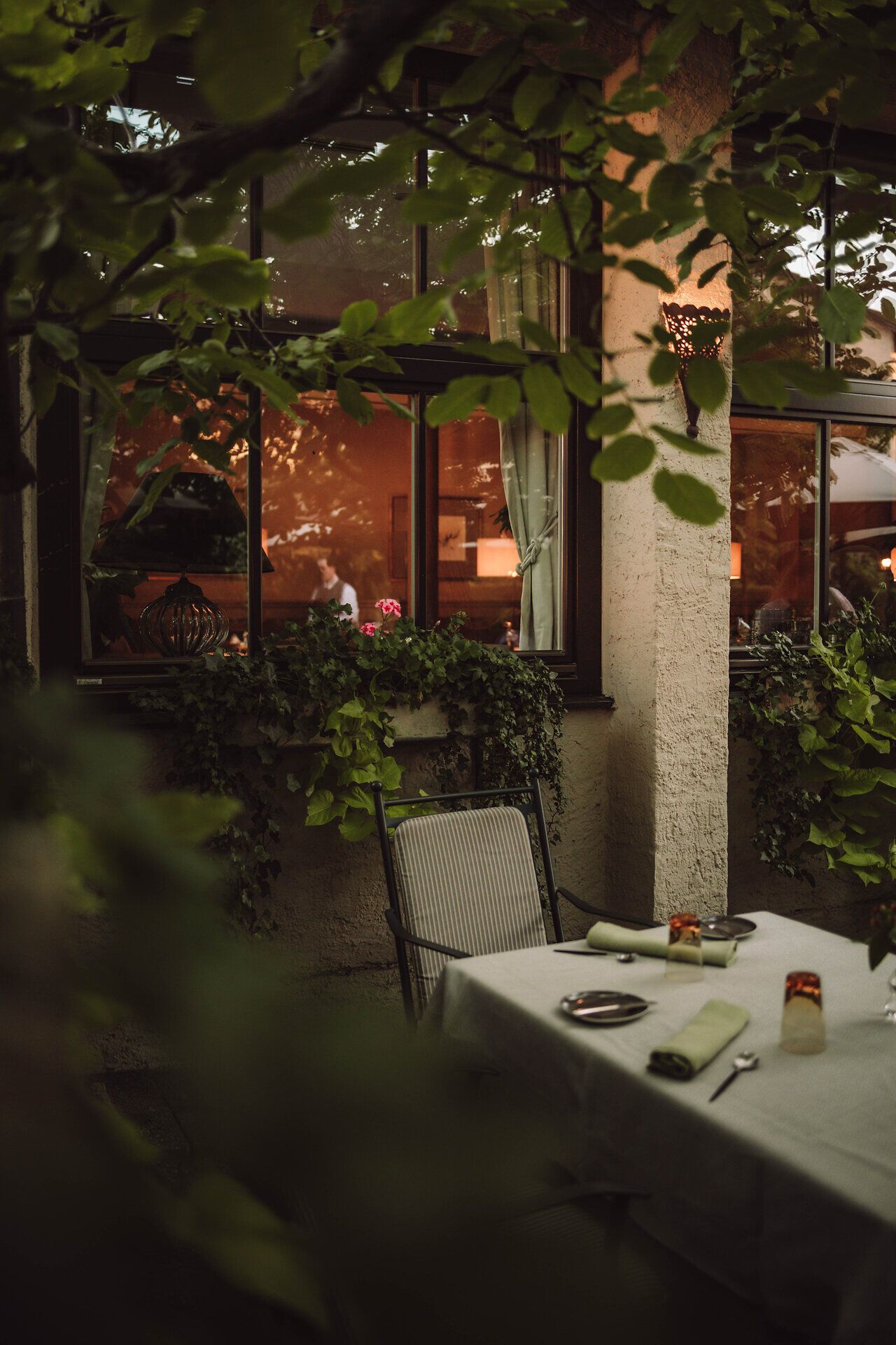 Ein einladender Tisch unter dem schattenspendenden Blätterdach lädt dazu ein, die köstlichen Spezialitäten der Region zu genießen. Die sanfte Abenddämmerung taucht die Umgebung in warmes Licht und schafft eine harmonische Atmosphäre, die zum Verweilen einlädt.