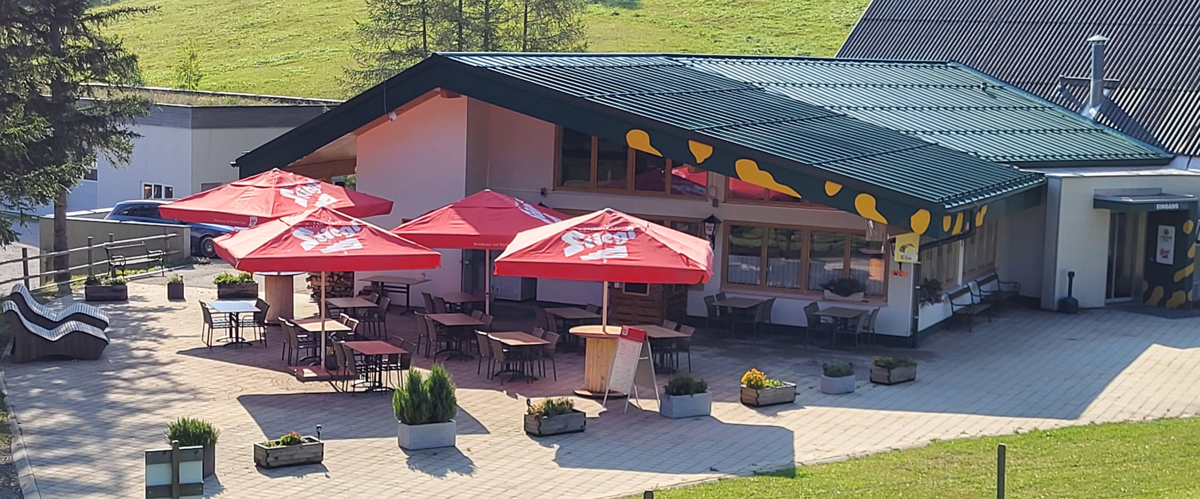 Außenansicht eines Restaurants mit roten Sonnenschirmen und Sitzplätzen im Freien.