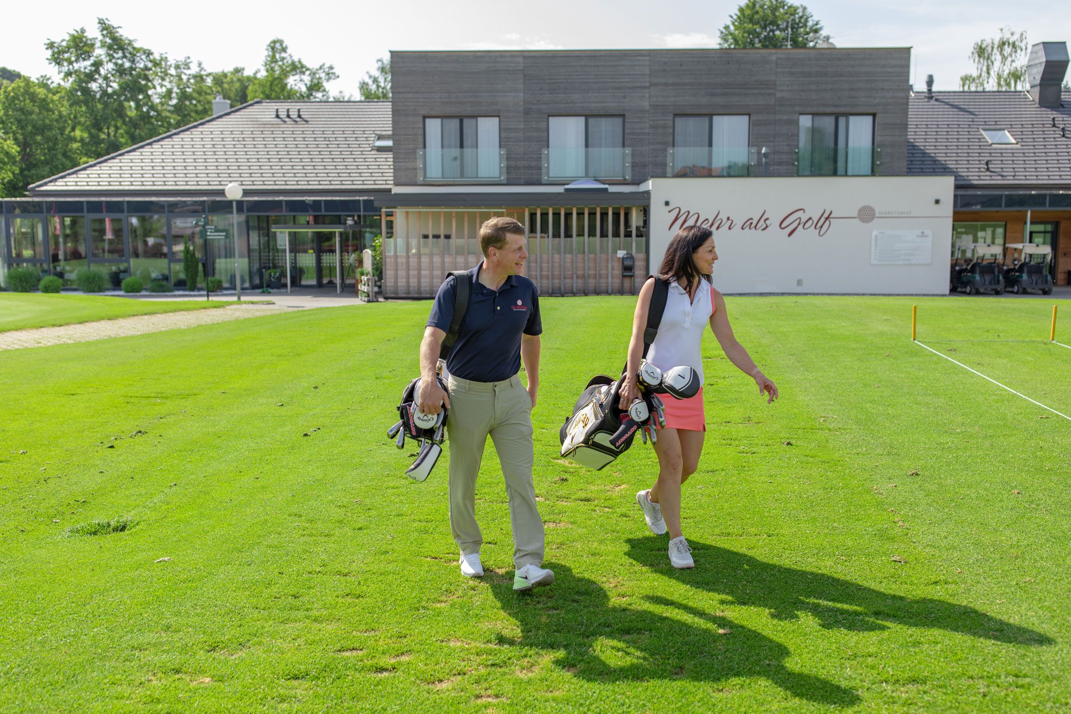 Zwei Personen mit Golftaschen vor einem modernen Clubhaus.