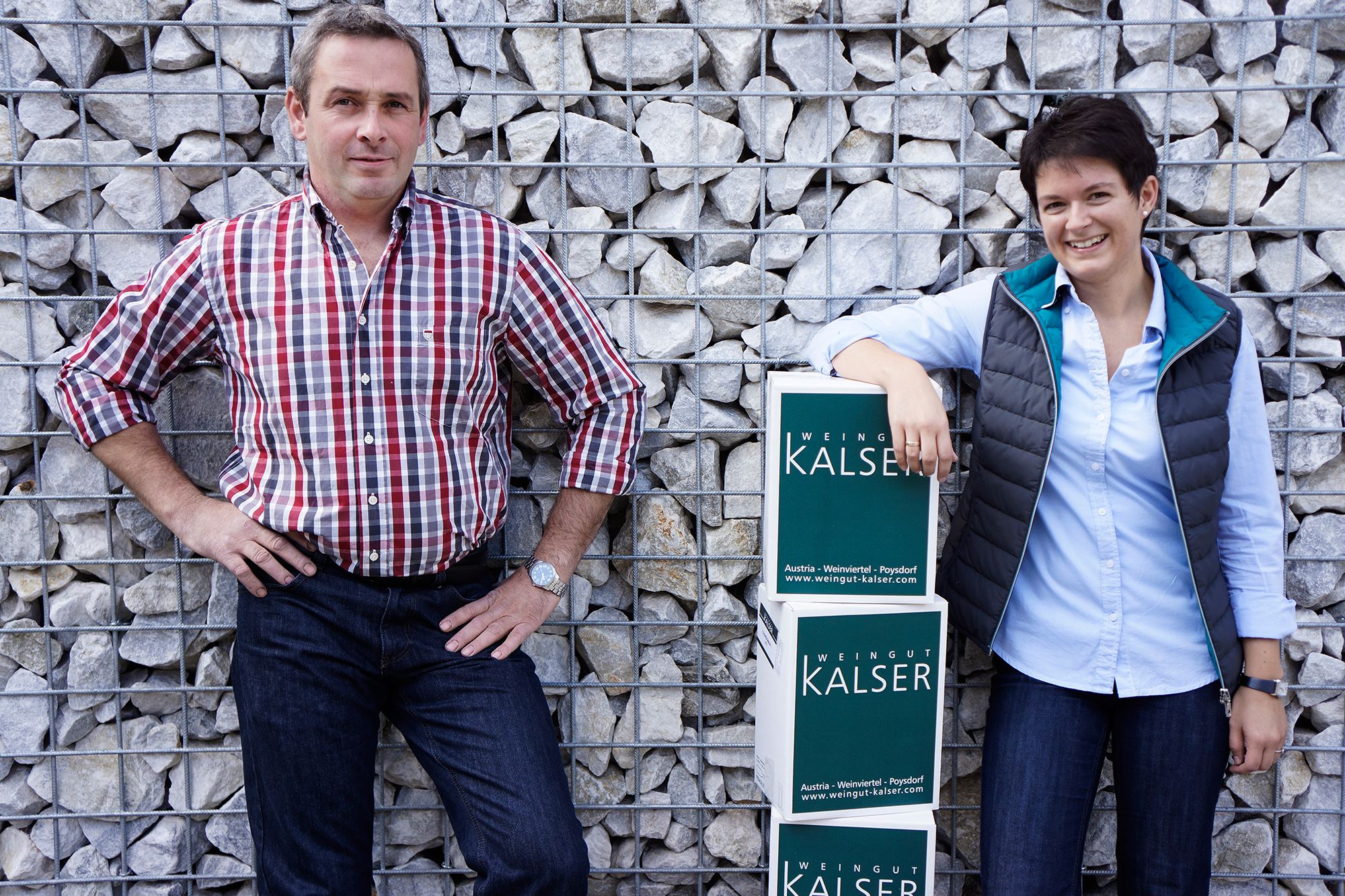 Zwei Personen stehen vor einer Steinmauer mit Kisten des Weinguts Kalser.