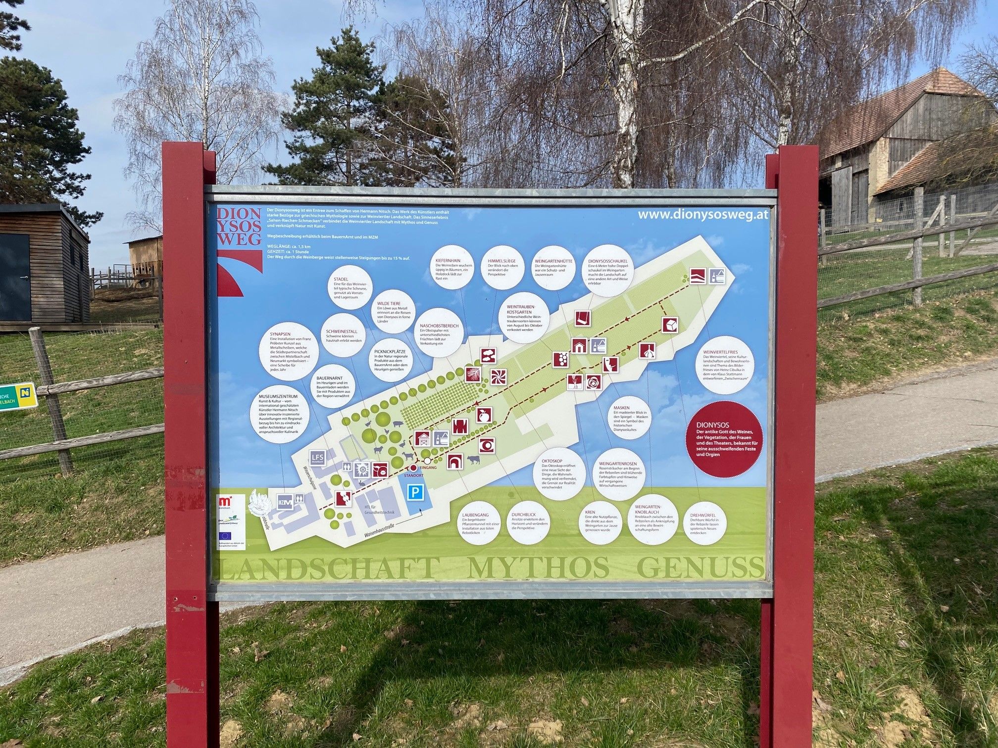 Informationsschild des Dionysoswegs mit Karte und Erklärungen, umgeben von Bäumen und Gebäuden.