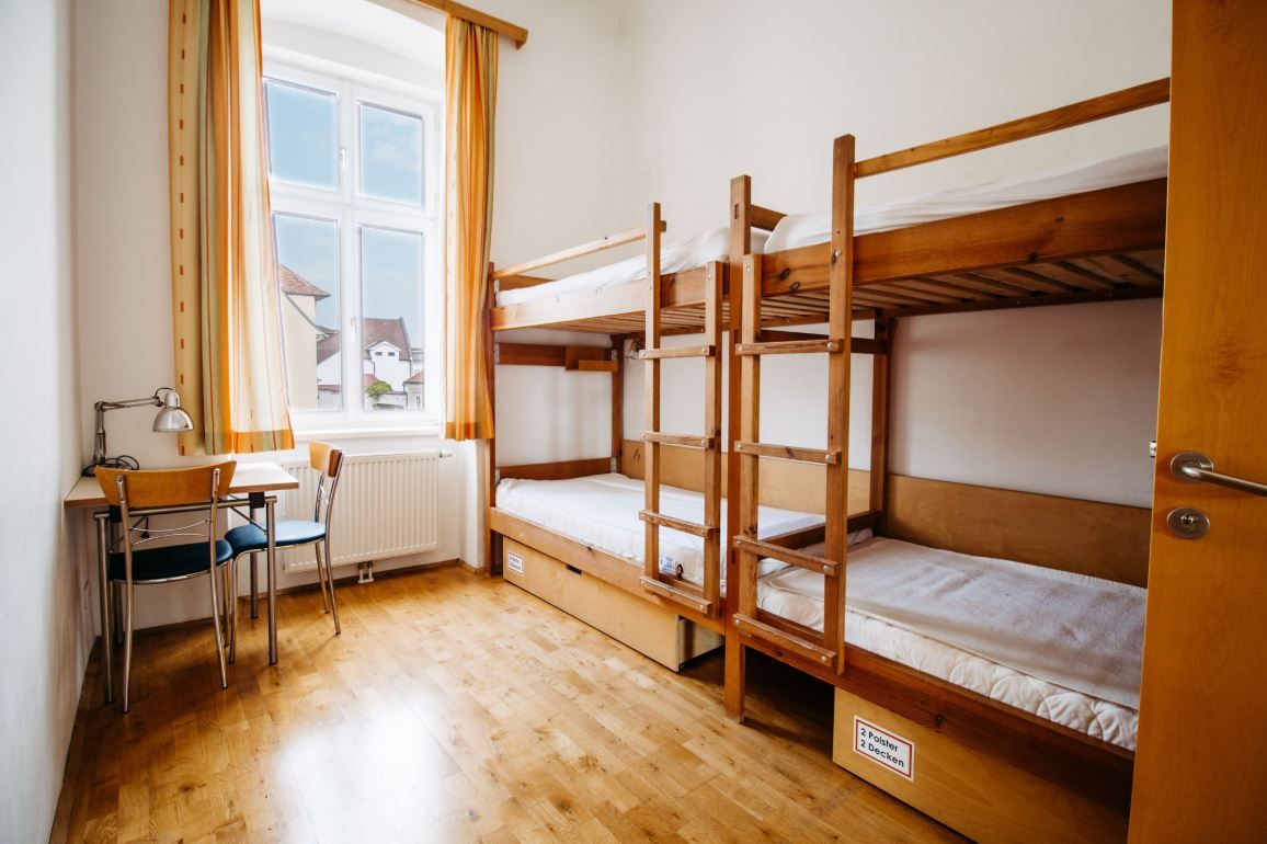 Ein einfaches Zimmer mit Etagenbett, Tisch und Stuhl, helle Vorhänge am Fenster.