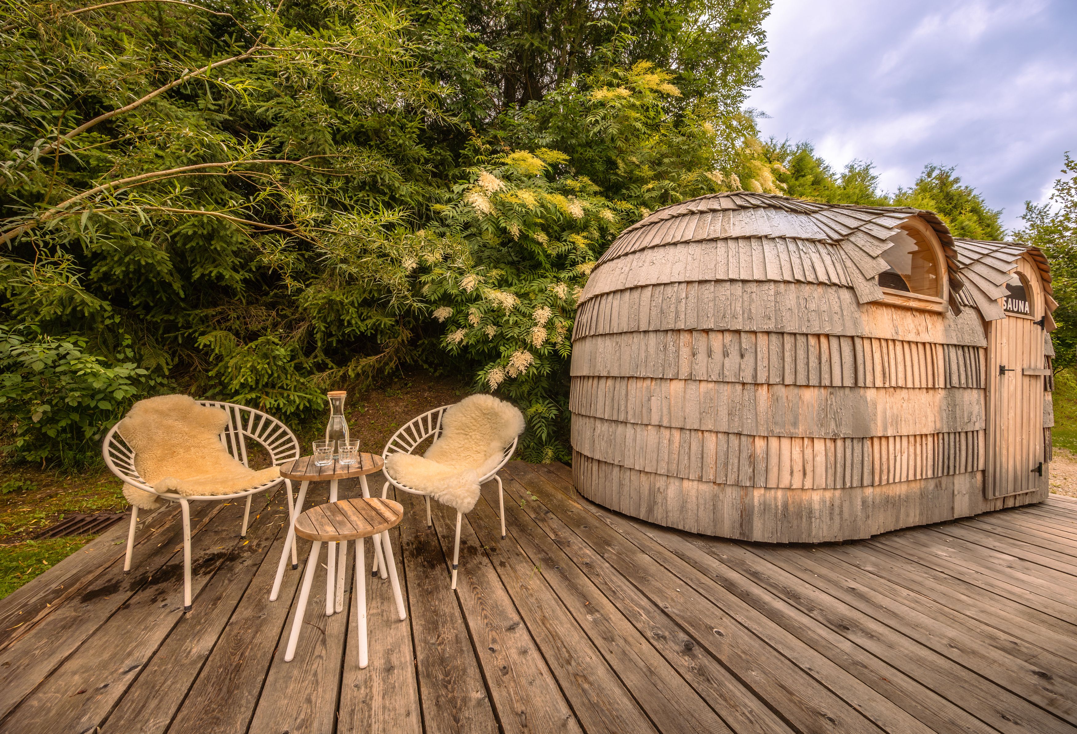 Holzsauna mit zwei Stühlen und Tisch auf einer Holzterrasse im Grünen.