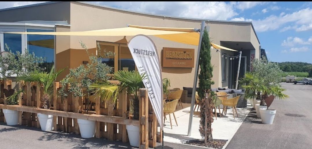 Außenansicht eines Cafés mit Terrasse, Sonnensegeln und Pflanzen.