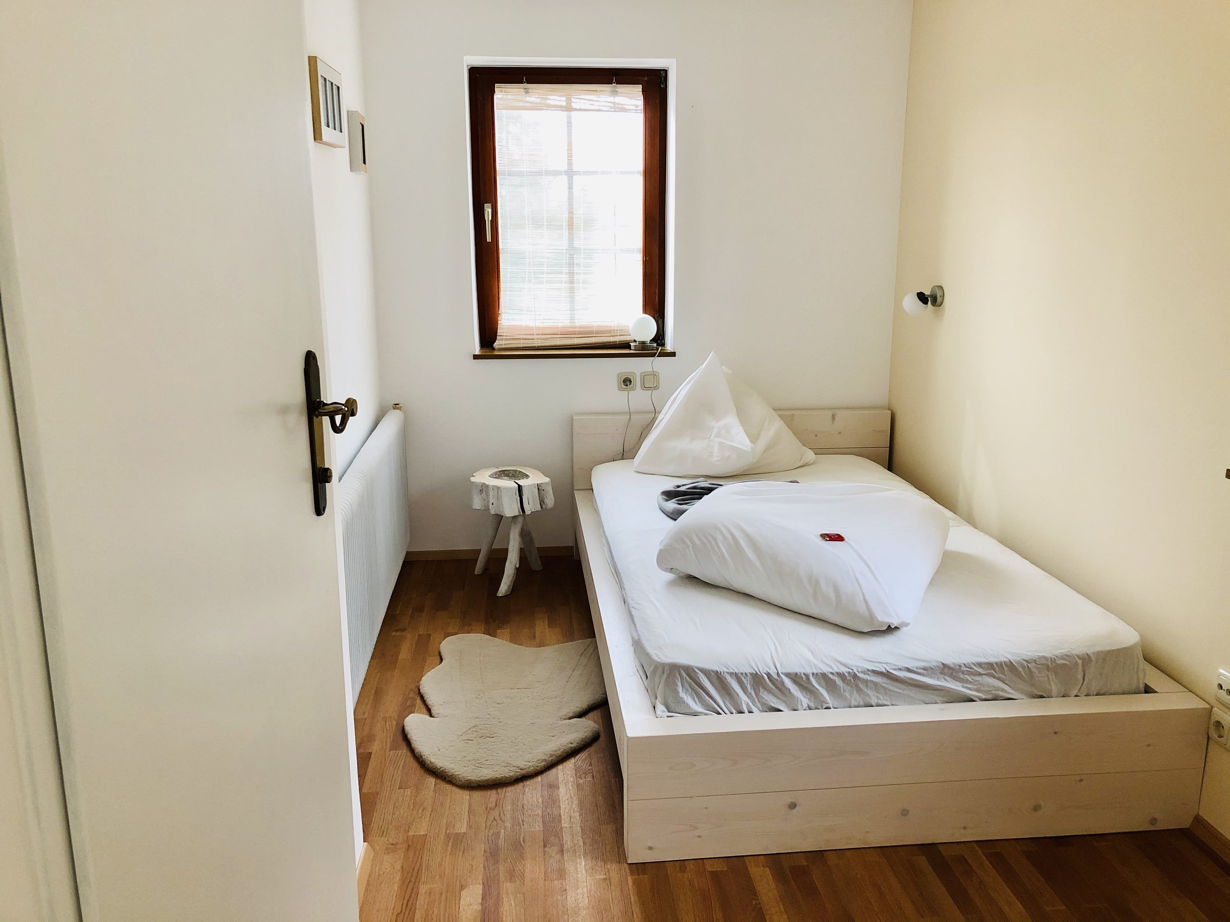 Ein kleines, gemütliches Schlafzimmer mit einem Einzelbett, Holzboden und einem kleinen Fenster mit Jalousie.