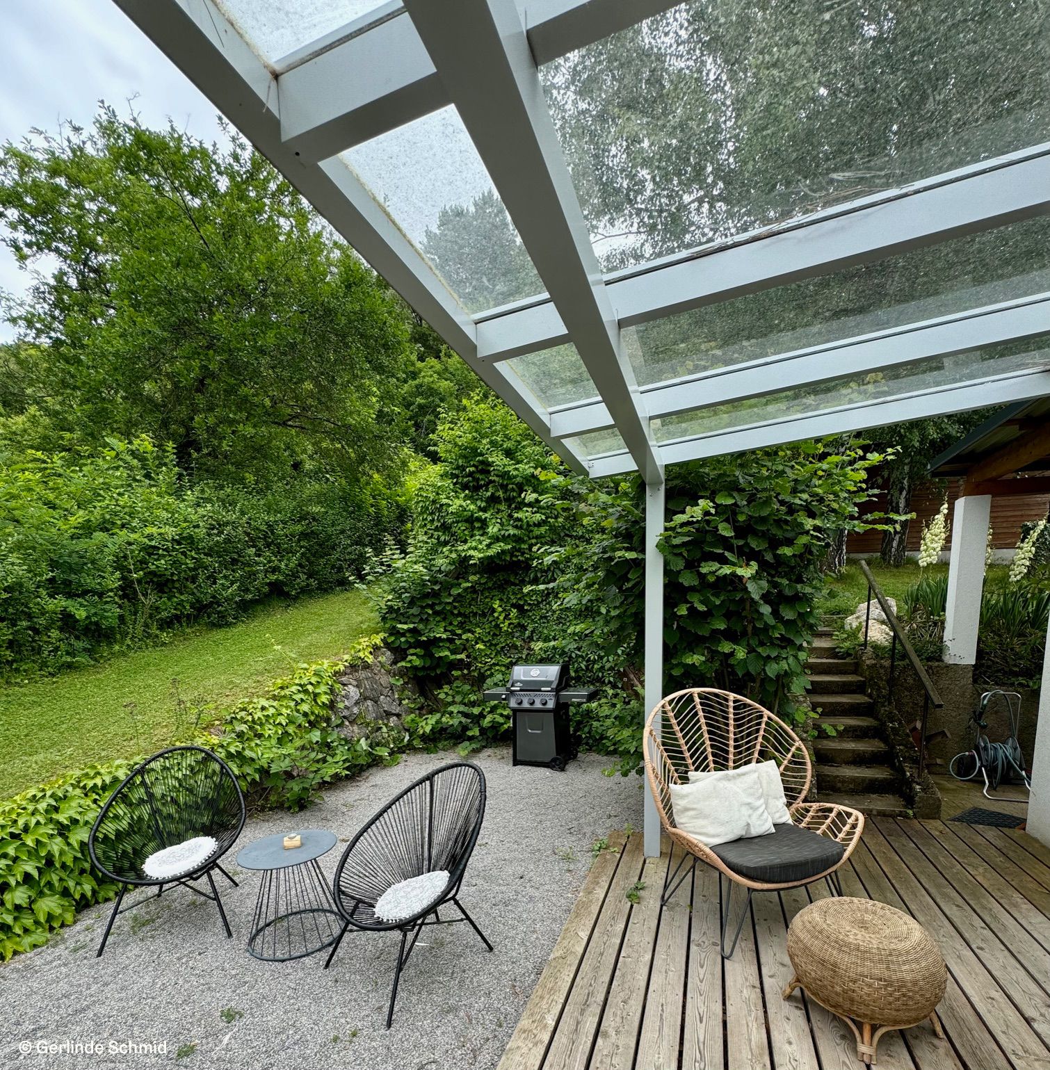 Terrasse mit Glasdach, Gartenmöbeln und Grill, umgeben von grüner Vegetation.