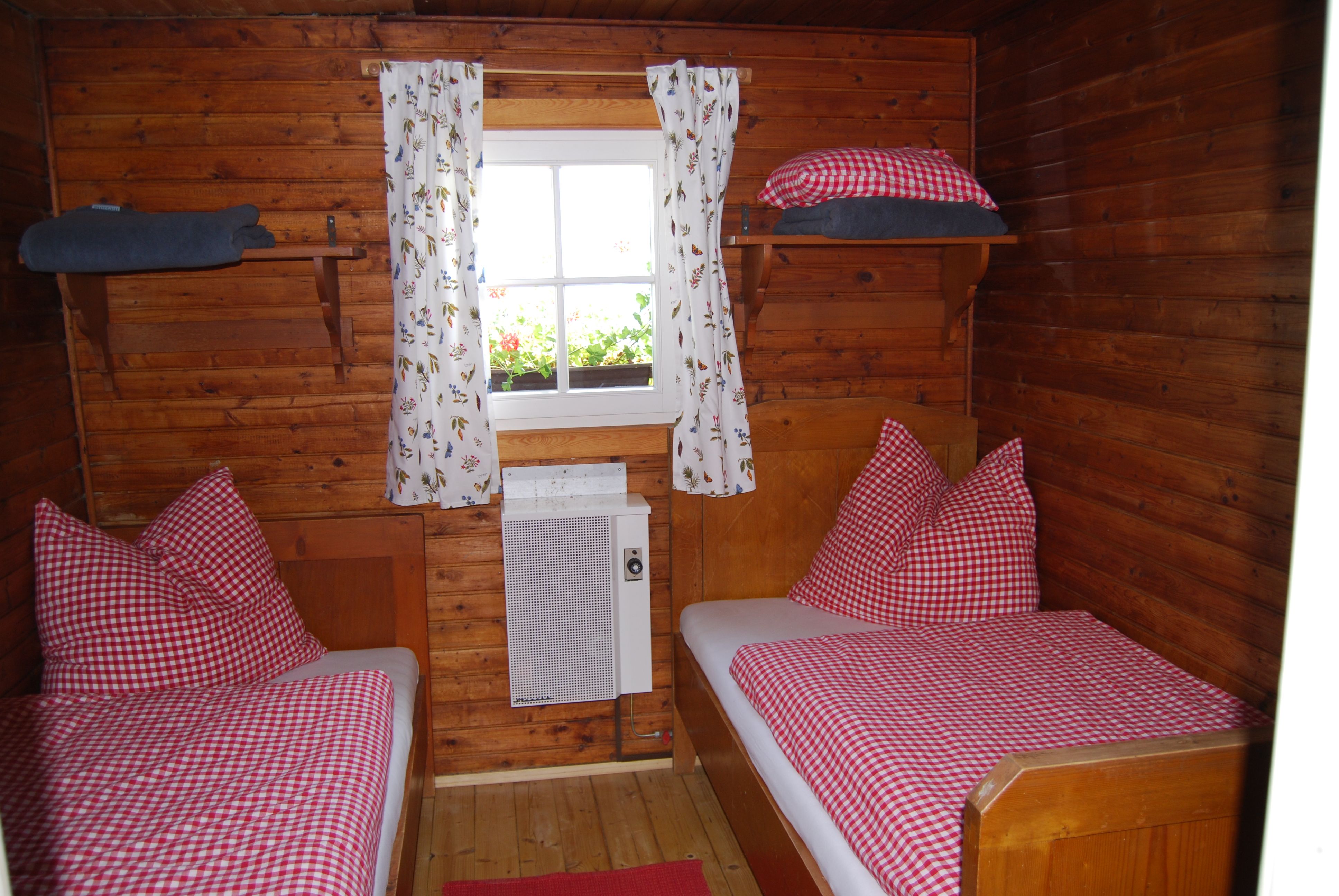Holzvertäfeltes Zimmer mit zwei Einzelbetten, rot-weiß karierten Bettdecken und einem Fenster mit Blumenvorhängen.