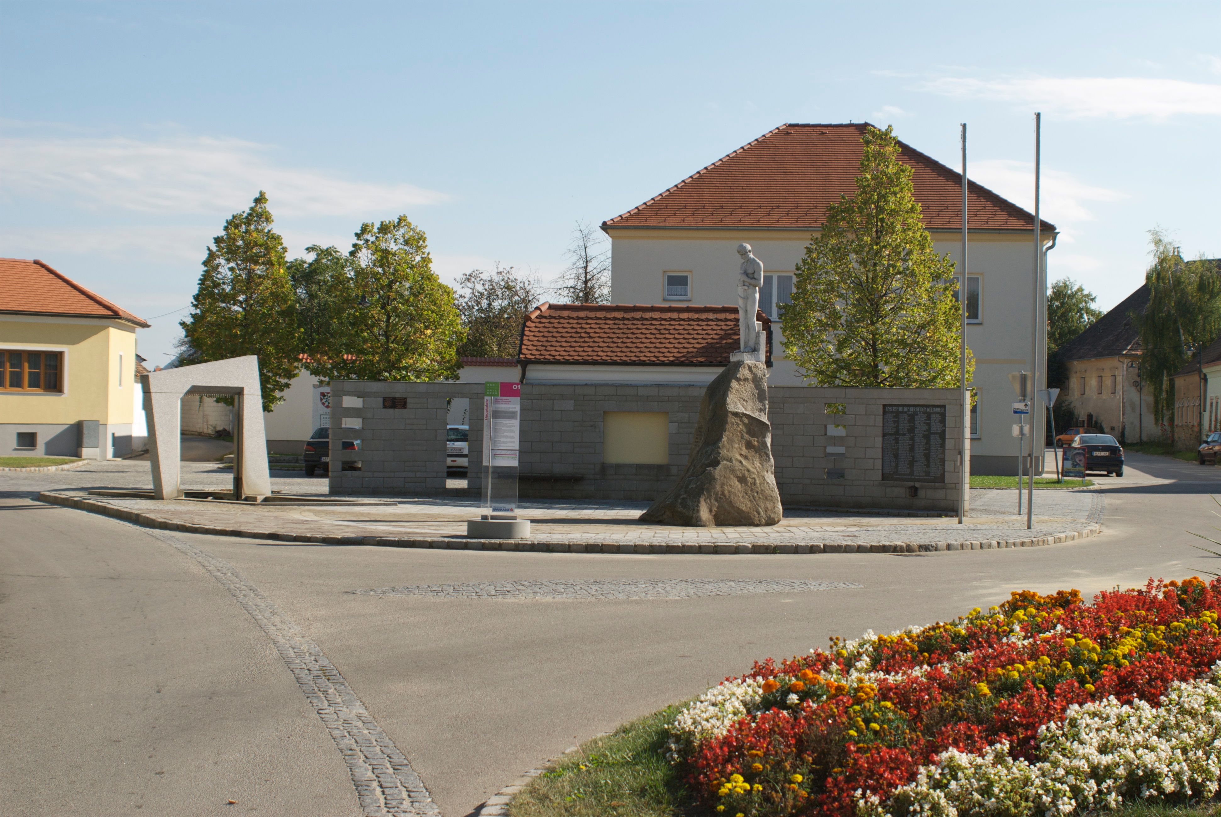 Denkmal in Mailberg mit Statue auf einem Felsen, umgeben von Gebäuden und Blumenbeeten.