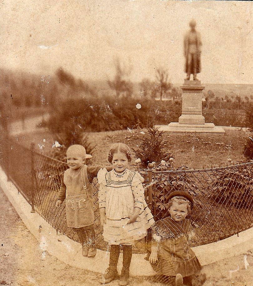 Drei Kinder stehen vor einer Statue in einem historischen Park.
