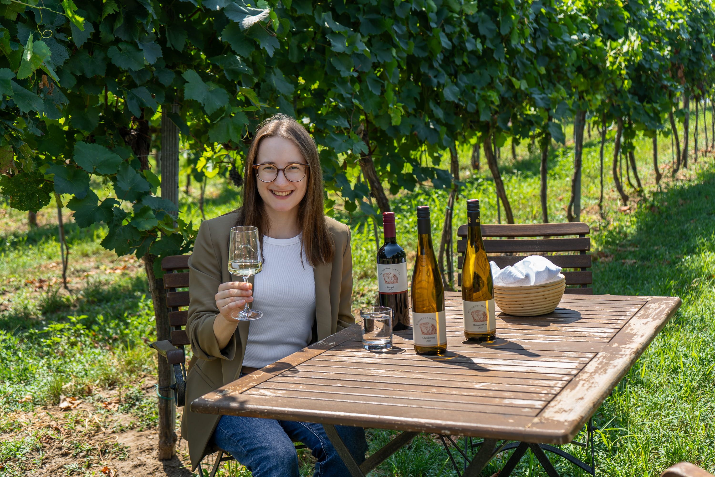 Eine Frau sitzt an einem Holztisch im Weinberg und hält ein Weinglas. Auf dem Tisch stehen mehrere Weinflaschen.