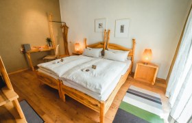 Ein gemütliches Schlafzimmer mit Holzmöbeln, einem Doppelbett, Nachttischen und einem Schreibtisch. Zwei Lampen und Bilder an der Wand ergänzen das Ambiente.