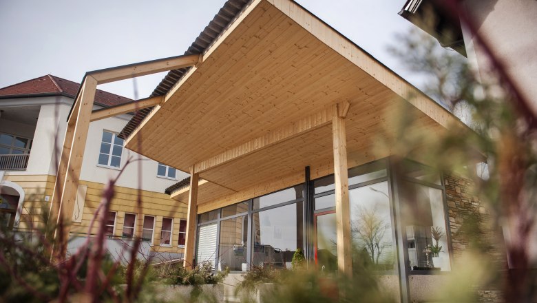 Modernes Geb&auml;ude mit Holz&uuml;berdachung und Glasfront, umgeben von Pflanzen.