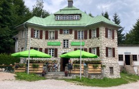 Steingebäude mit grünem Dach und Sonnenschirmen vor dem Eingang.