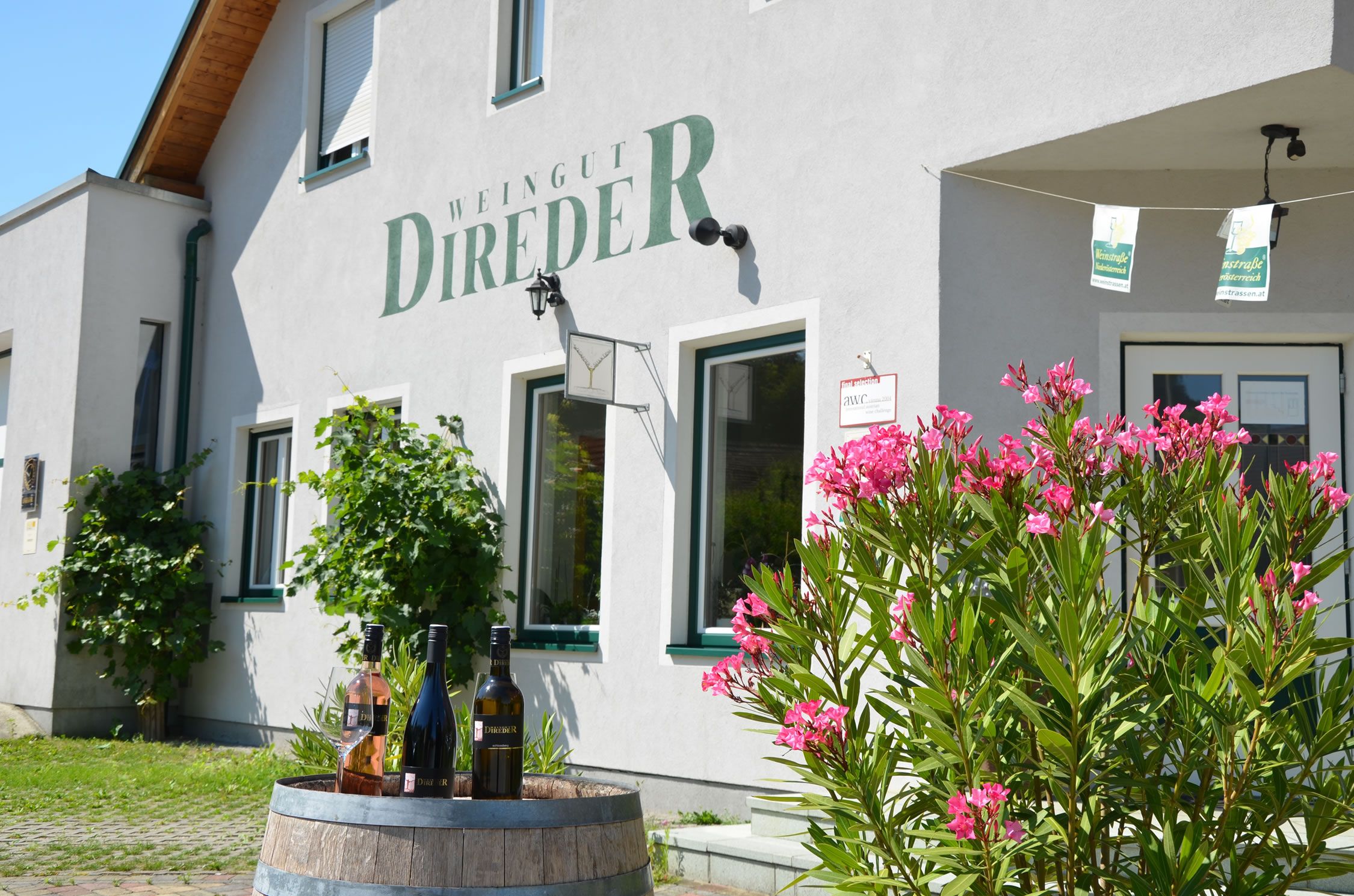 Fassade eines Weinguts mit Weinflaschen auf einem Fass und blühendem Oleander im Vordergrund.