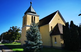 Gelbe Kapelle mit Turm und Kreuz, umgeben von B&auml;umen, unter blauem Himmel.