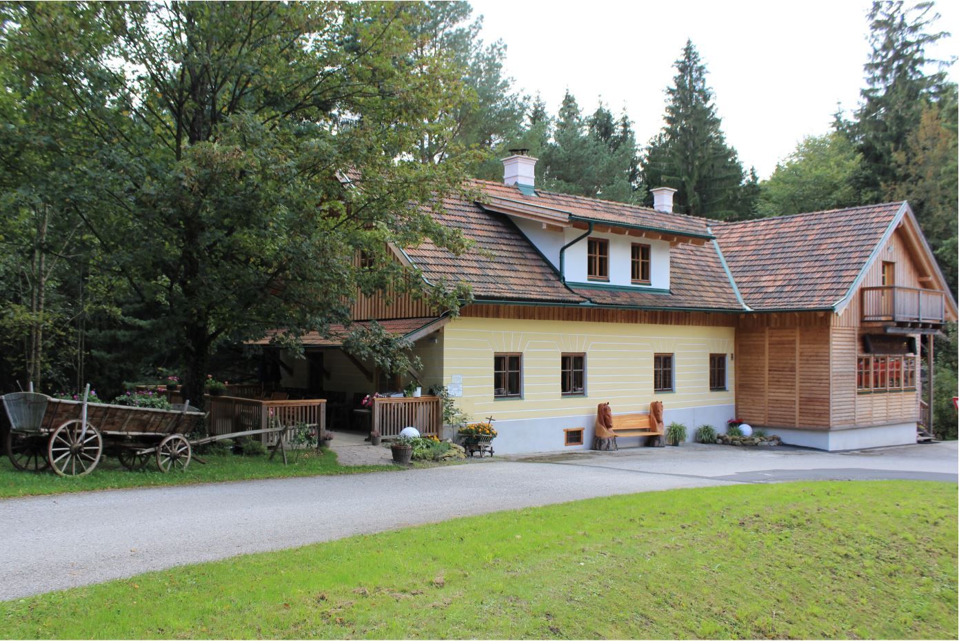 Ein traditionelles Gasthaus mit Holzdach und gelber Fassade, umgeben von Bäumen und einem alten Holzwagen im Vordergrund.