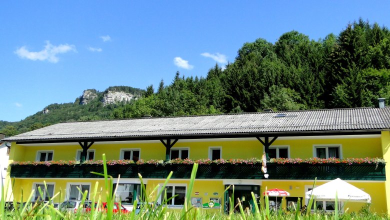 Voralpenhof, © Voralpenhof Gelbes Gebäude mit Blumenbalkon vor bewaldetem Hügel und blauem Himmel.