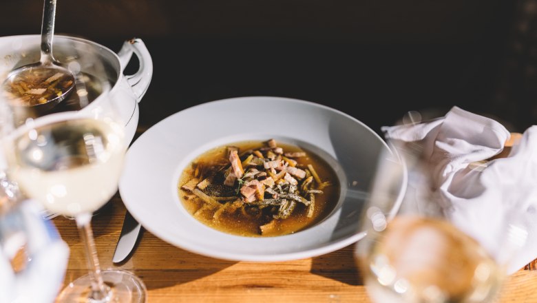Ein Teller mit Roggenbiersuppe und Mohnfrittaten auf einem Holztisch, daneben ein Glas Wei&szlig;wein.