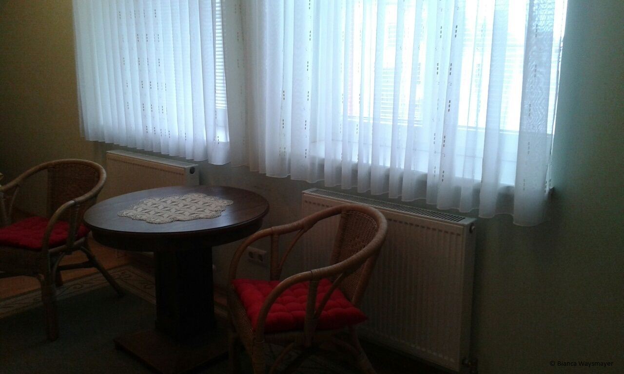 Zwei Korbstühle mit roten Kissen und ein runder Holztisch vor einem Fenster mit weißen Vorhängen.