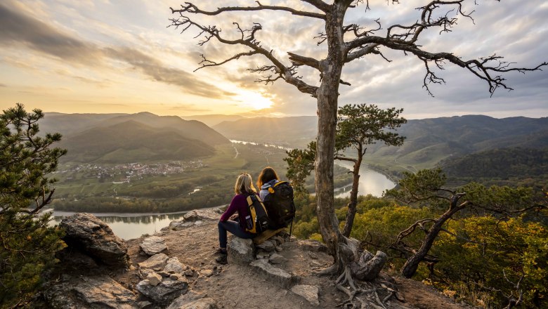 Zwei Personen sitzen auf einem Felsen und blicken auf eine Flusslandschaft bei Sonnenuntergang.