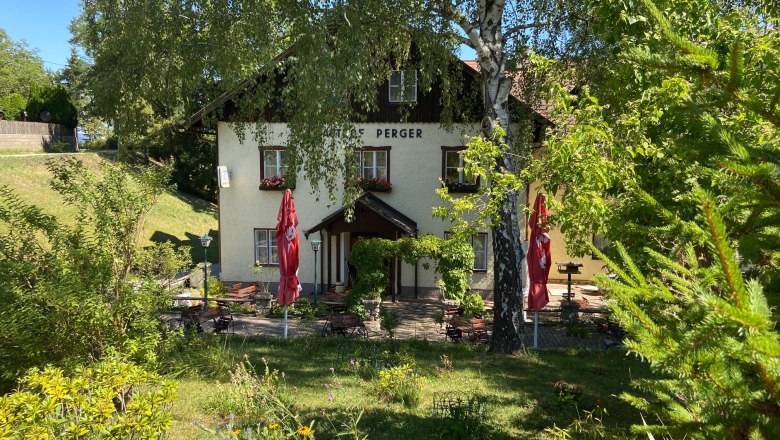 Ein Gasthaus mit Terrasse, umgeben von B&auml;umen und Pflanzen, bei sonnigem Wetter.