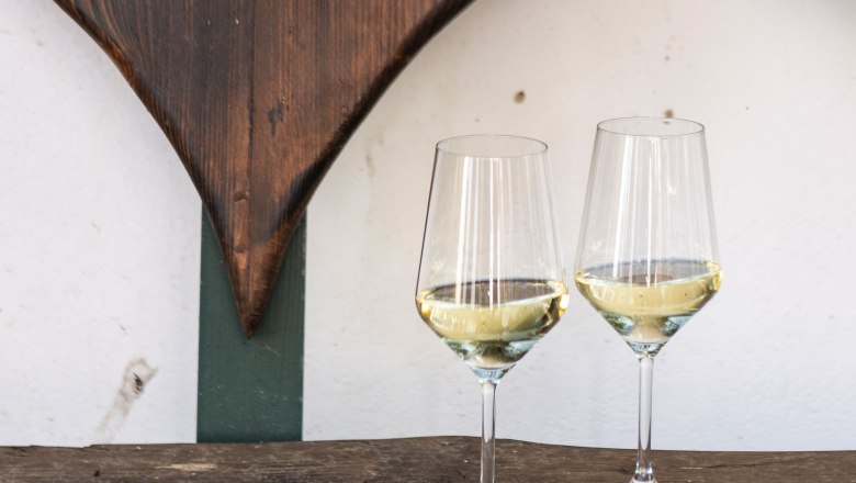 Zwei Weingl&auml;ser mit Wei&szlig;wein auf einem Holztisch vor einer Holzherz-Dekoration.