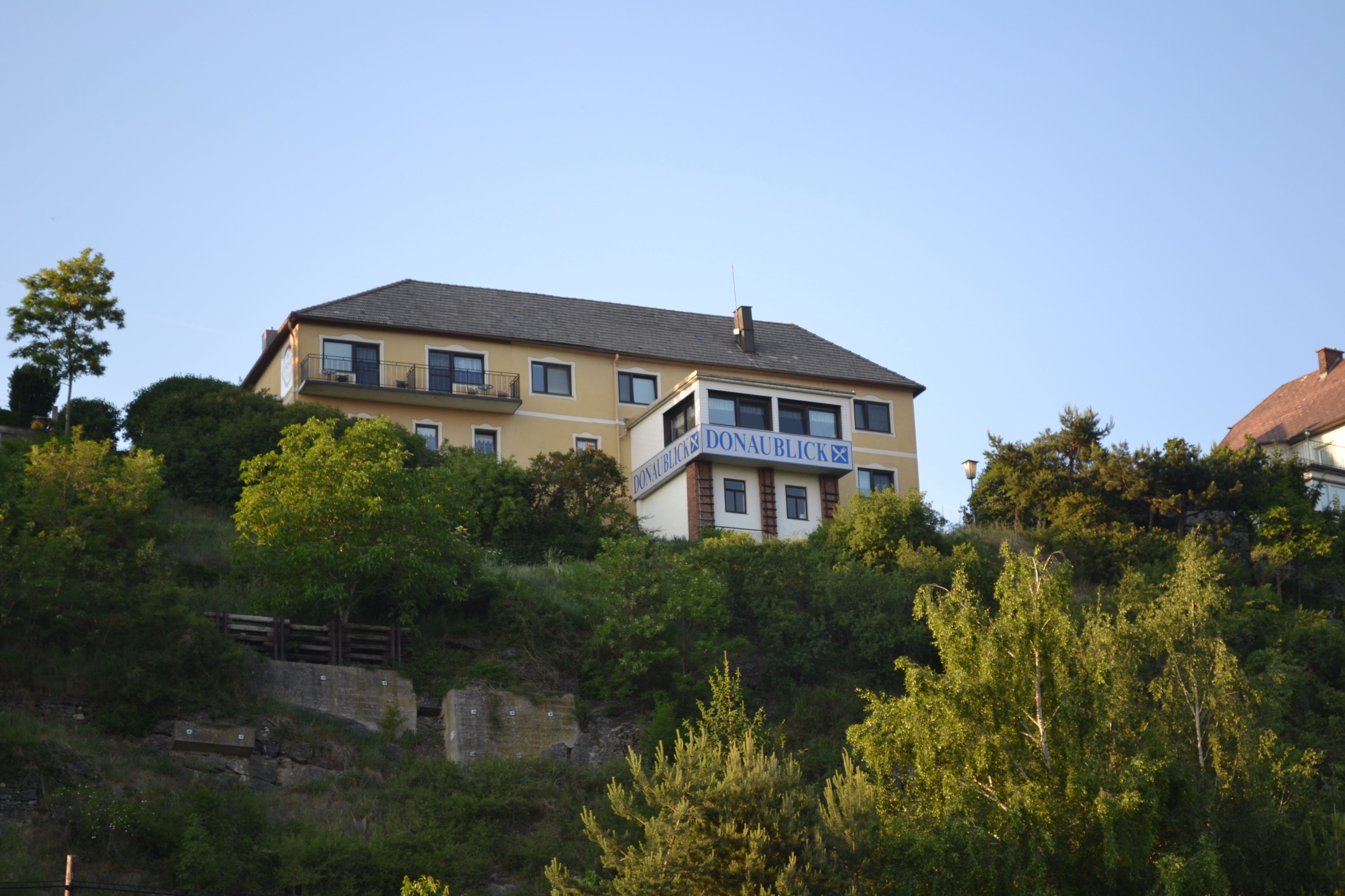Ein gelbes Gebäude mit der Aufschrift 'Donaublick' auf einem Hügel, umgeben von Bäumen und blauem Himmel.