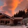 Ein Holzchalet im Schnee bei Sonnenuntergang mit orangefarbenem Himmel und B&auml;umen im Hintergrund.