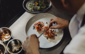 Koch dekoriert ein Gericht mit Lachs und Gem&uuml;se auf einem Teller.