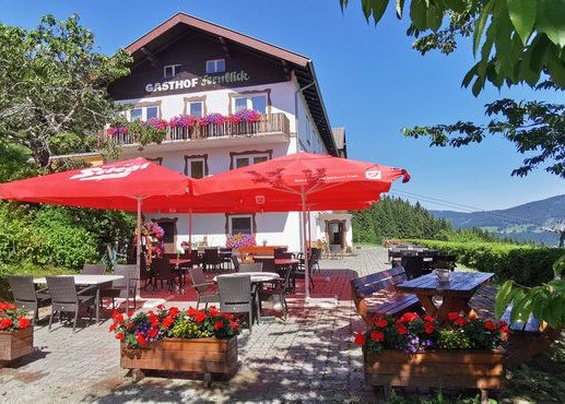 Ein traditioneller Gasthof mit Terrasse, roten Sonnenschirmen und Blumenk&auml;sten vor einem klaren blauen Himmel.
