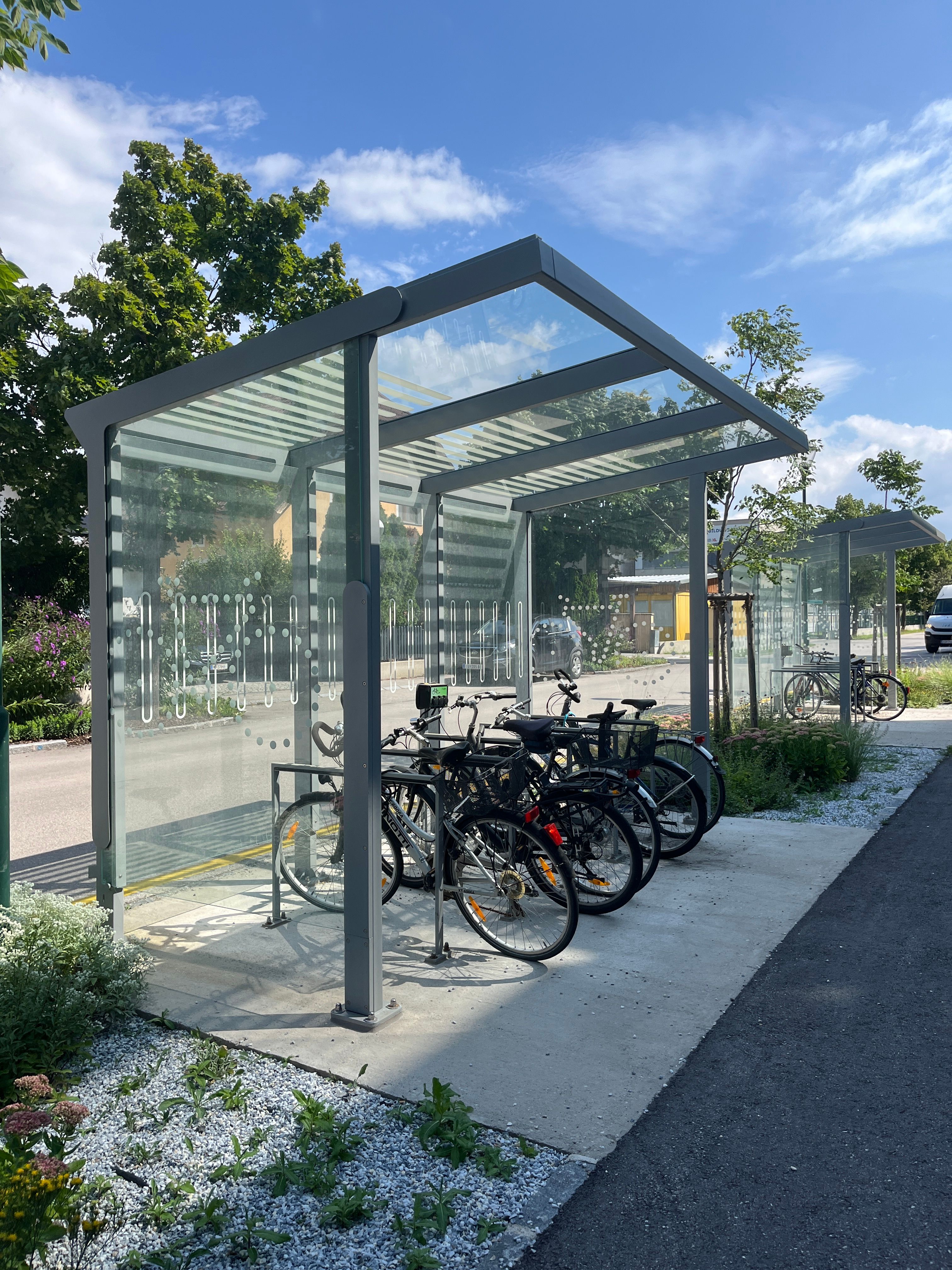 Überdachte E-Bike-Ladestation mit mehreren Fahrrädern, umgeben von Bäumen und Pflanzen.
