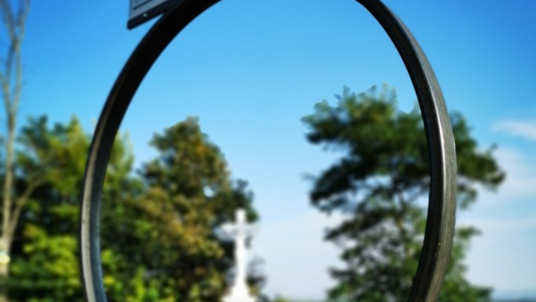 Ein Schild mit der Aufschrift 'Seefeld-Kadolz' vor einem blauen Himmel, im Hintergrund ein weißes Kreuz und Bäume.