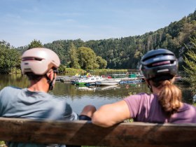 Die sanfte Brise streicht &uuml;ber den glitzernden Stausee, w&auml;hrend Radfahrer auf einer Bank entspannen und die malerische Landschaft genie&szlig;en. Umgeben von &uuml;ppigem Gr&uuml;n und sanften H&uuml;geln, l&auml;dt dieser Ort dazu ein, die Seele baumeln zu lassen und die Sch&ouml;nheit der Natur zu erleben.