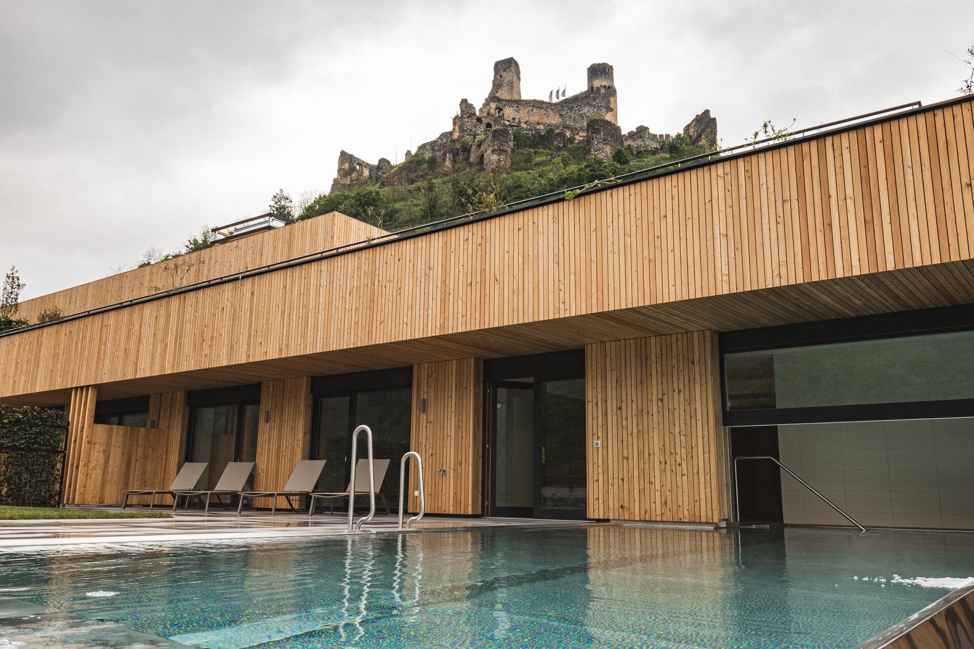 Moderner Außenpool mit Holzverkleidung und Burgruine im Hintergrund.