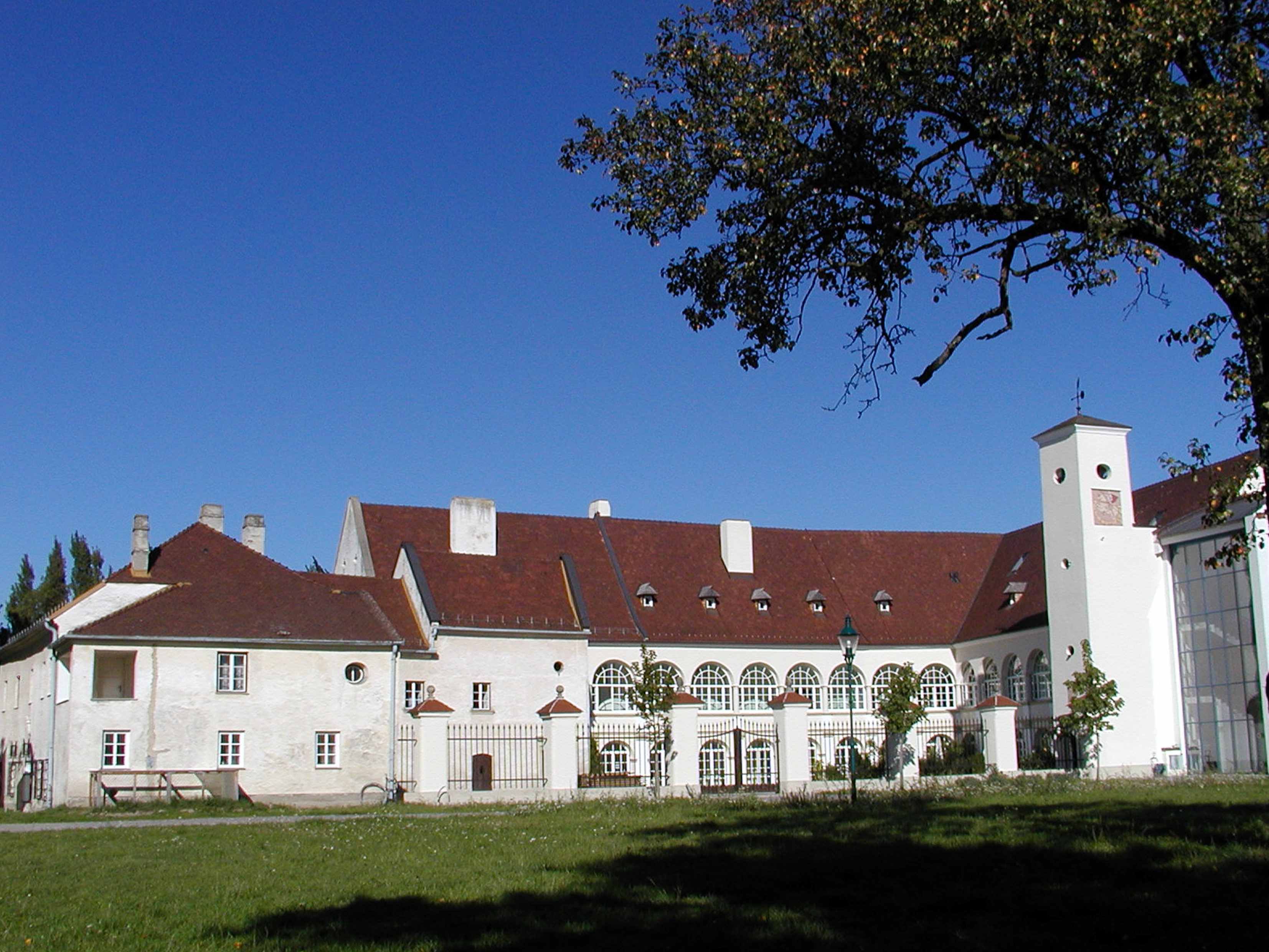 Schloss Katzelsdorf mit rotem Dach und weißen Wänden, umgeben von Bäumen und Rasen.