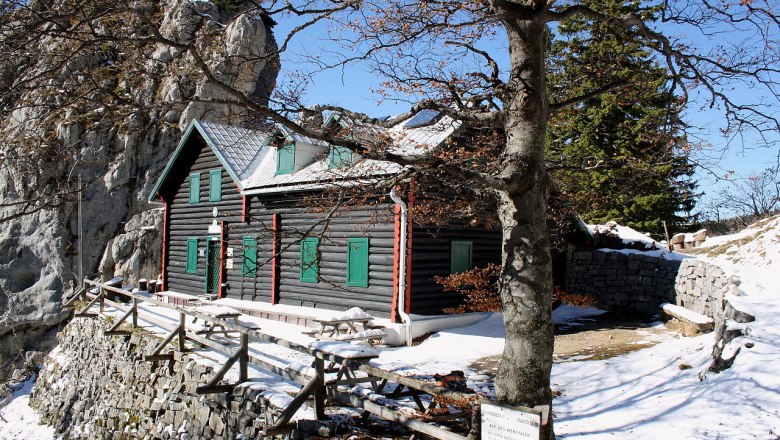 Eine Berghütte aus Holz mit grünen Fensterläden, umgeben von Schnee und Felsen, unter einem klaren blauen Himmel.