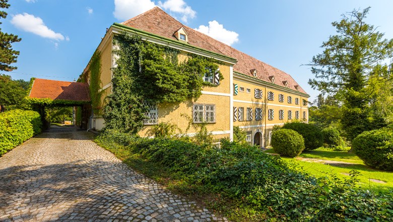 Ein historisches Schloss mit gelber Fassade, bewachsen mit Efeu, umgeben von gepflegtem Garten und gepflastertem Weg.