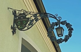 Metallschild mit Laterne an einer Hauswand, beschriftet mit 'Heuriger Fam. Almst&auml;dter'.