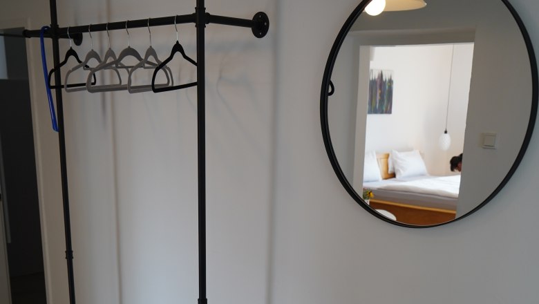 Innenansicht eines Zimmers mit Kleiderständer, rundem Spiegel und kleinem Tisch mit Pflanze und Vase.