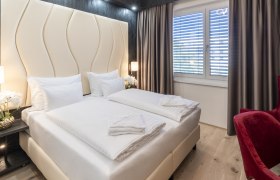 Modernes Hotelzimmer mit Doppelbett, roten Sesseln und gro&szlig;en Fenstern mit Vorh&auml;ngen.
