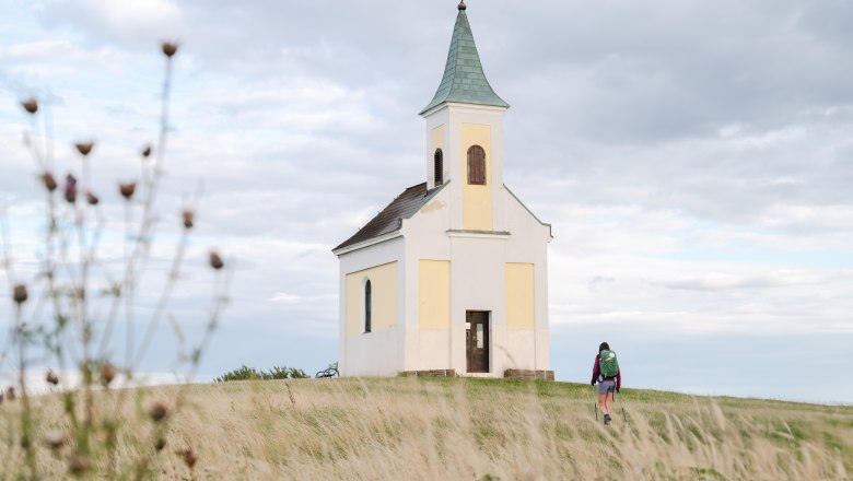 Eine kleine Kapelle auf einem H&uuml;gel mit einer Person, die darauf zugeht, umgeben von einer Wiese und bew&ouml;lktem Himmel.
