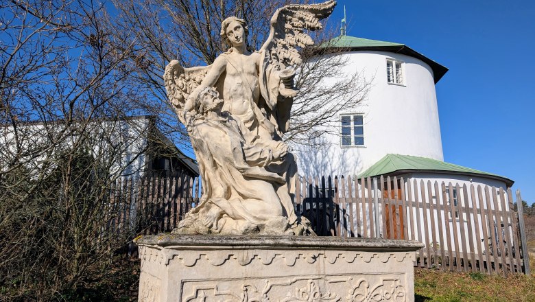 Steinstatue eines Engels mit Fl&uuml;geln und einer knienden Figur vor einem runden, wei&szlig;en Geb&auml;ude mit gr&uuml;nem Dach.