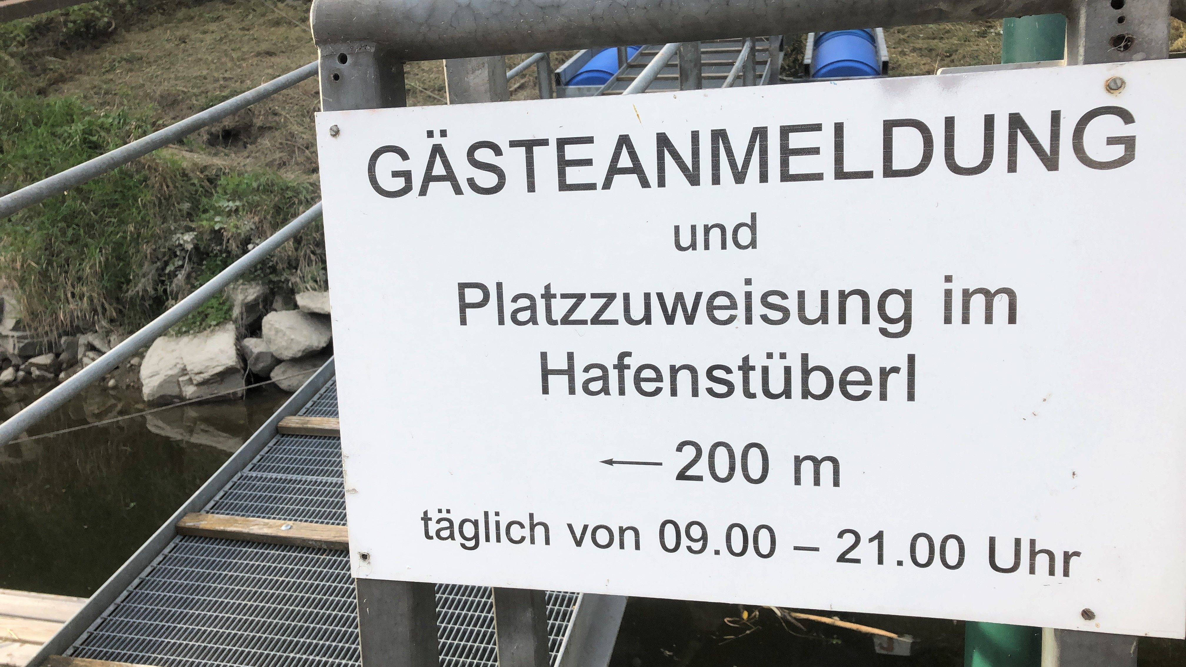 Ein Schild mit der Aufschrift 'Gästeanmeldung und Platzzuweisung im Hafenstüberl, 200 m, täglich von 09.00 – 21.00 Uhr'.