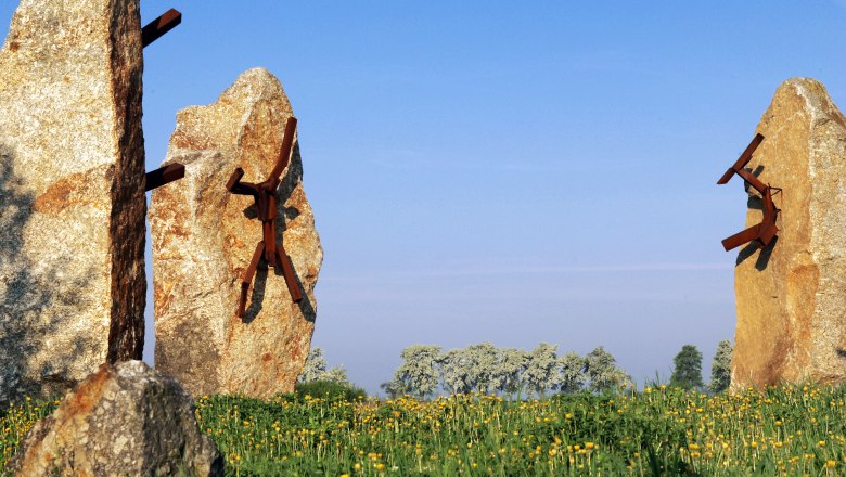 Steinskulpturen mit Metallfiguren auf einer Wiese unter blauem Himmel.